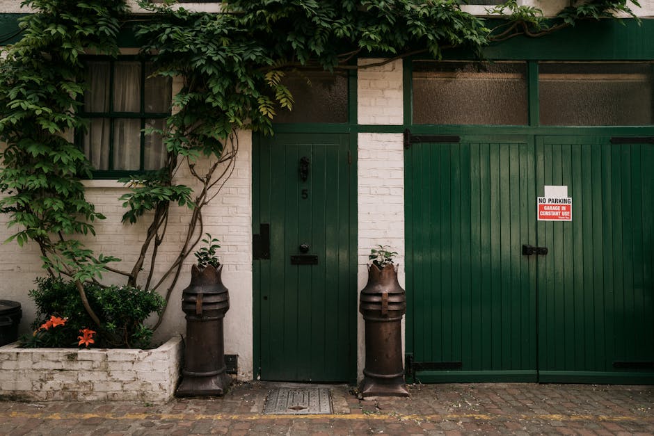 A close-up view of a charming garden alleyway adjacent to a white brick building, featuring a dark green wooden door with the number 5, and a large sliding green garage door. Flanking the door are two