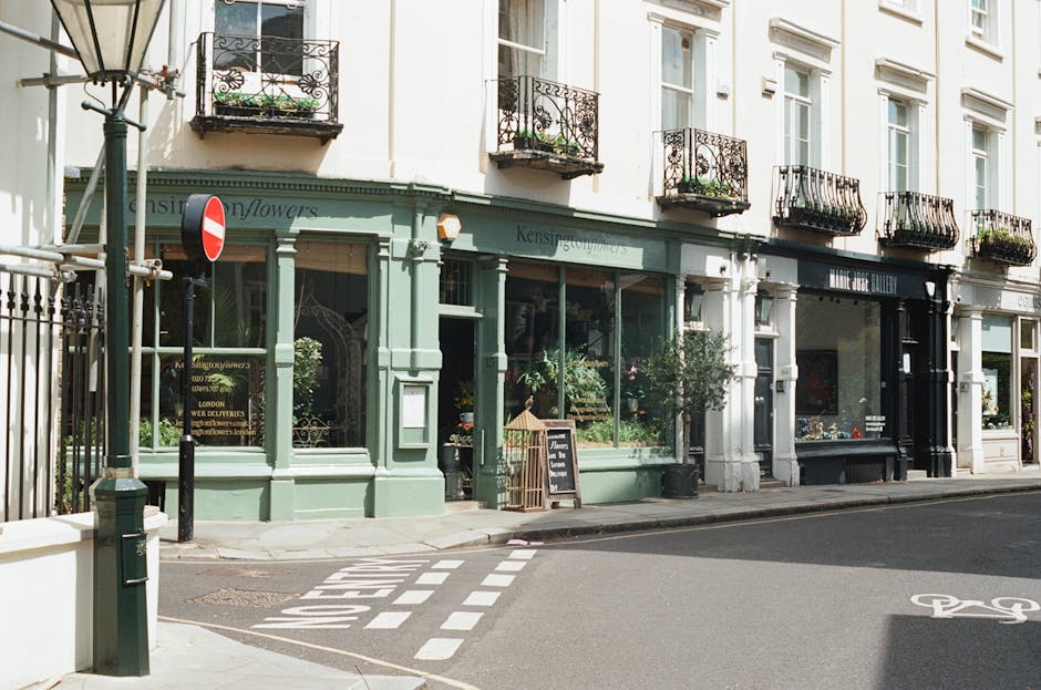 A close-up view of a charming garden alleyway adjacent to a white brick building, featuring a dark green wooden door with the number 5, and a large sliding green garage door. Flanking the door are two